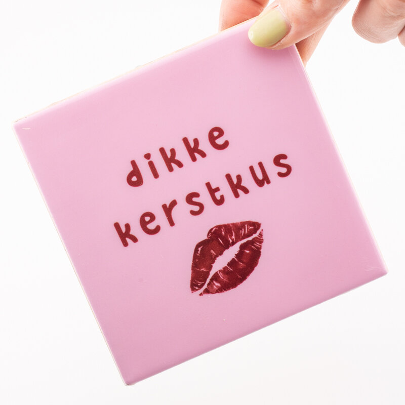 Hand houdt roze tegeltje met tekst &quot;Dikke Kerstkus&quot; en lippenafdruk vast.